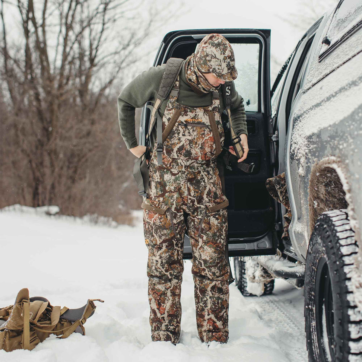 A person wears First Lite multi-color camouflage hunting bibs with padded shoulder straps in a snowy forest.