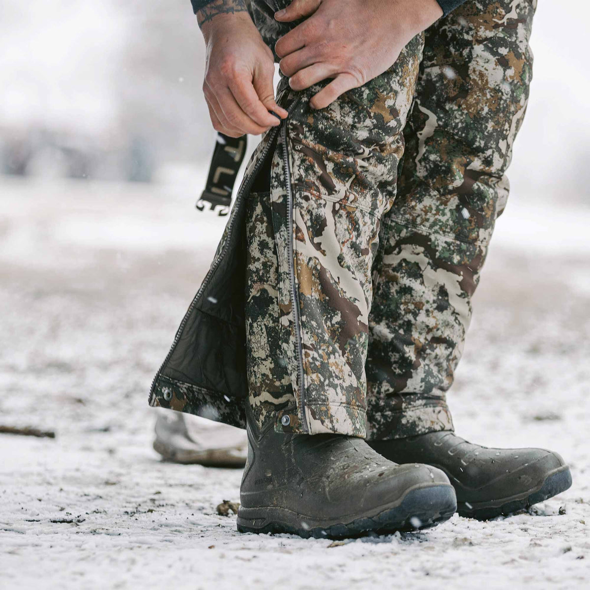 A person zips up camouflage pants with full-length side zippers over dark boots on snowy ground.