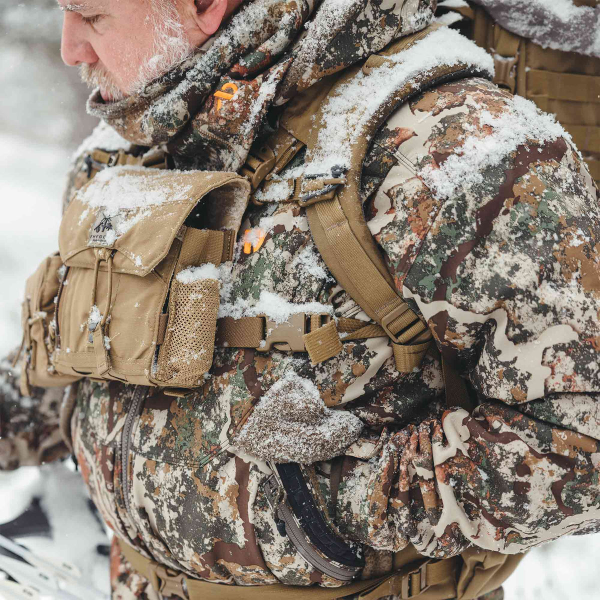 Snow-covered multi-color camouflage hunting jacket worn with a tan chest rig featuring a flap closure and mesh pocket.