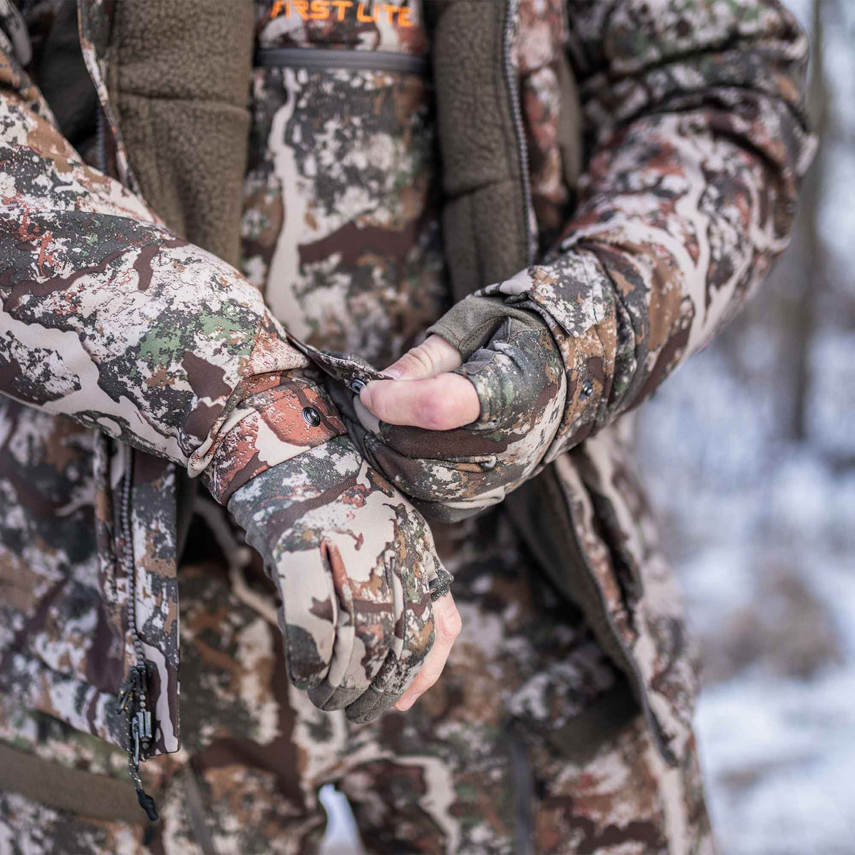 A person in a brown, green, tan, and white camouflage hunting jacket adjusts the snap-cuff over matching fingerless gloves.