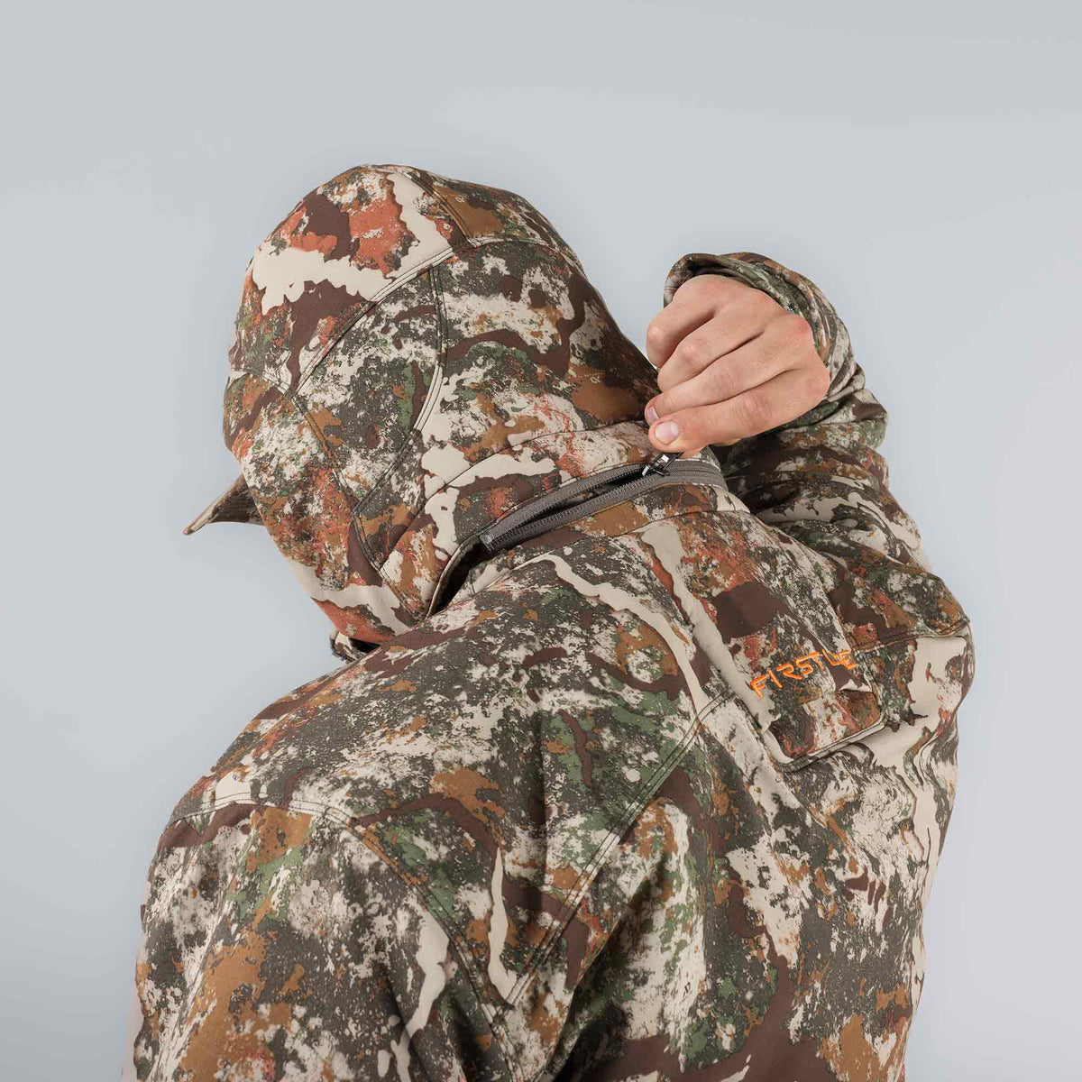 Camouflage hunting jacket with hood up, a person zipping a grey diagonal chest pocket, and an orange "FIRST" logo visible on the garment.