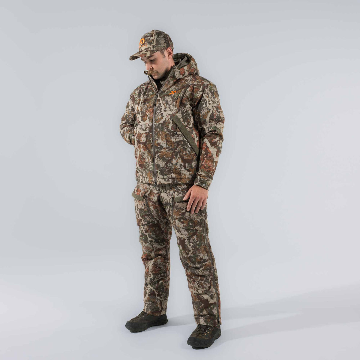 Men's camouflage hunting suit with a hooded jacket featuring an orange logo, matching pants with side zippers, and a mesh-back cap.