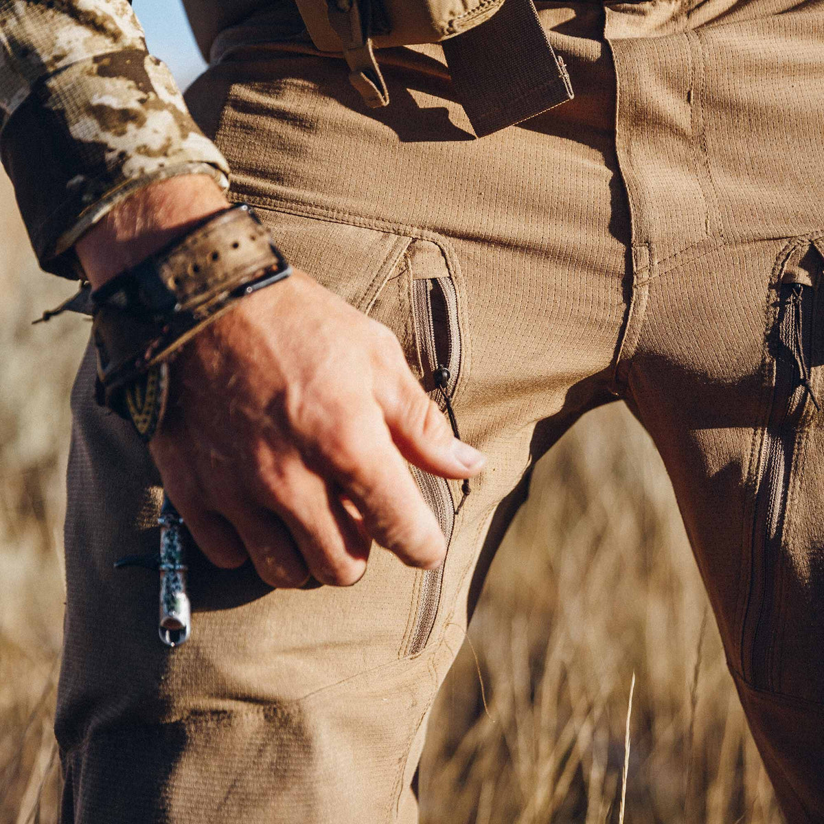Tan tactical pants with a vertically textured fabric and zippered side pockets, worn by a person holding a small tool.