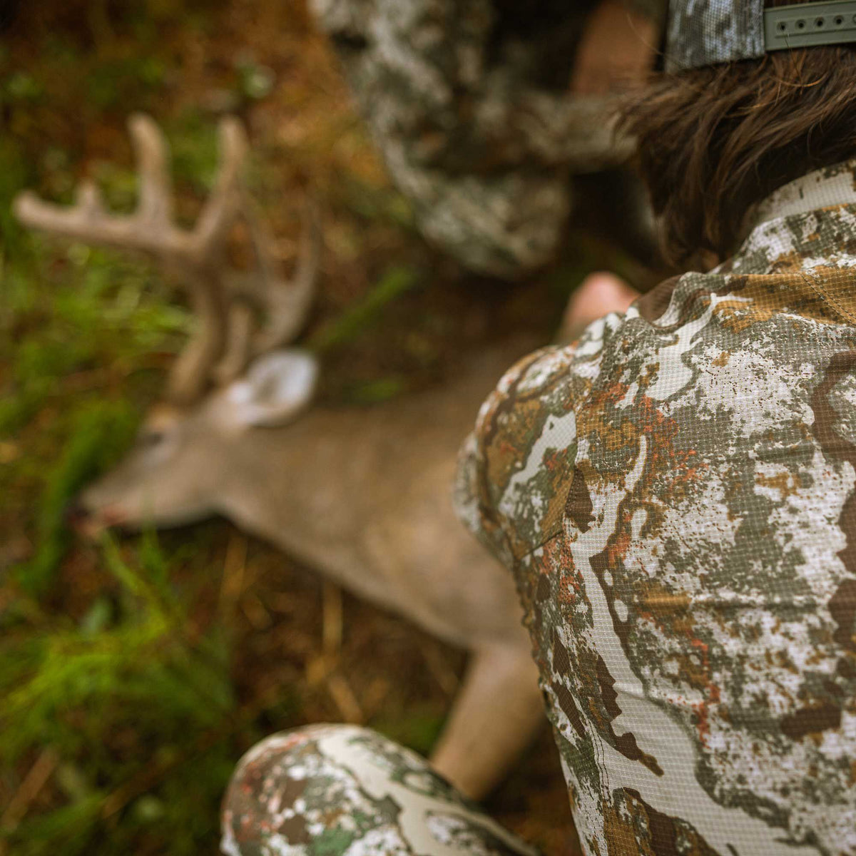 Rear view of a person wearing a textured earth-toned camouflage hunting shirt with a ribbed collar and snapback hat, kneeling by a deer.