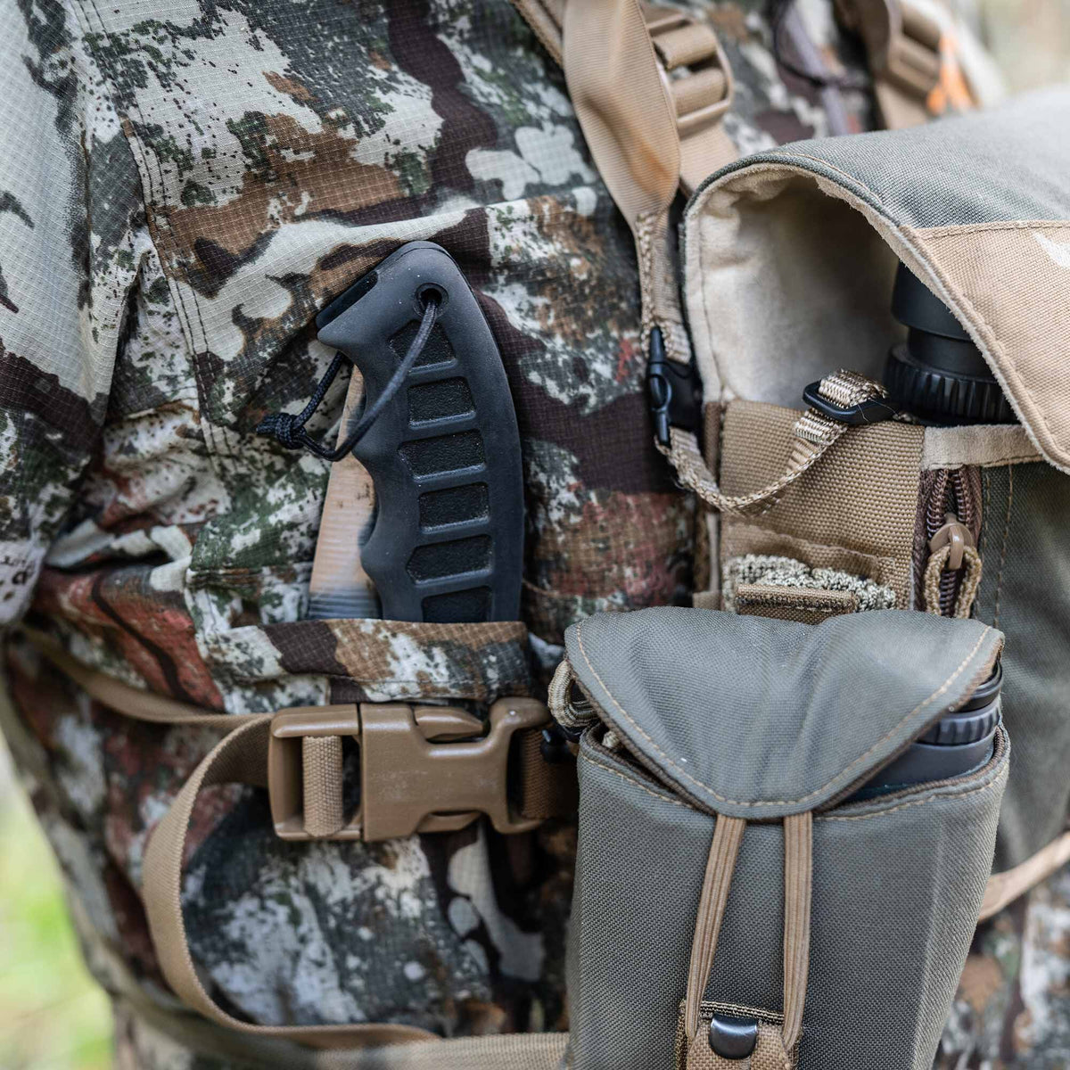 An olive green chest rig is worn over camouflage clothing, featuring a black tool handle and black binoculars visible in front pouches.