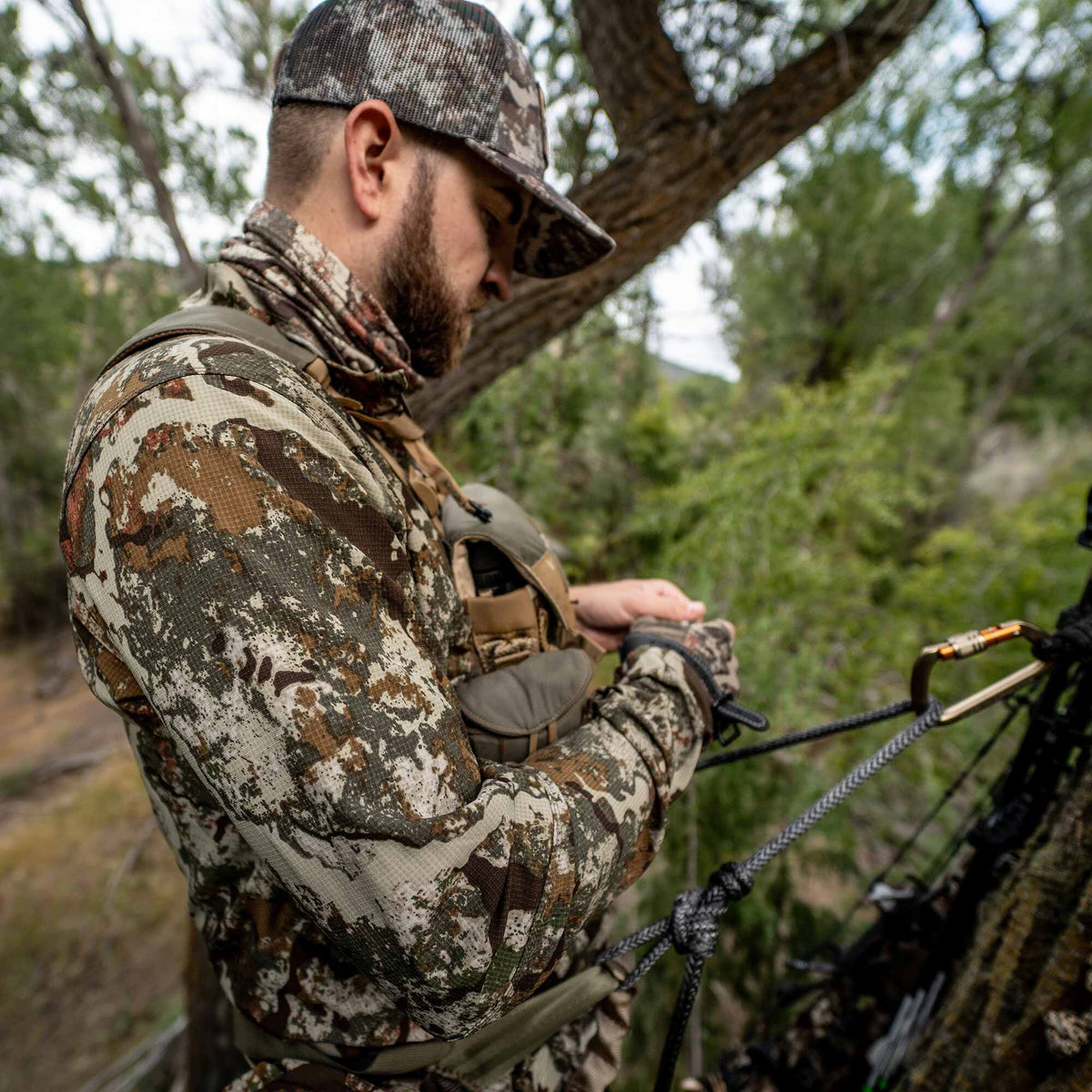 A hunter wears a multi-color camouflage long-sleeve shirt, neck gaiter, and cap, with a chest harness holding binoculars, while adjusting a rope on a bow setup.