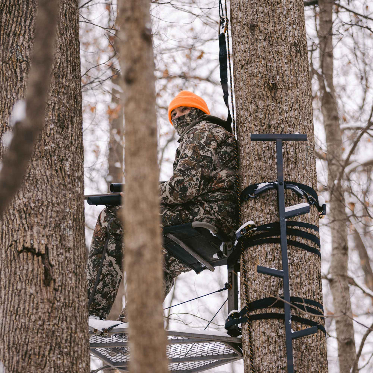 Man sitting in a chair tied to a tree and wearing camo jacket and camo pants.