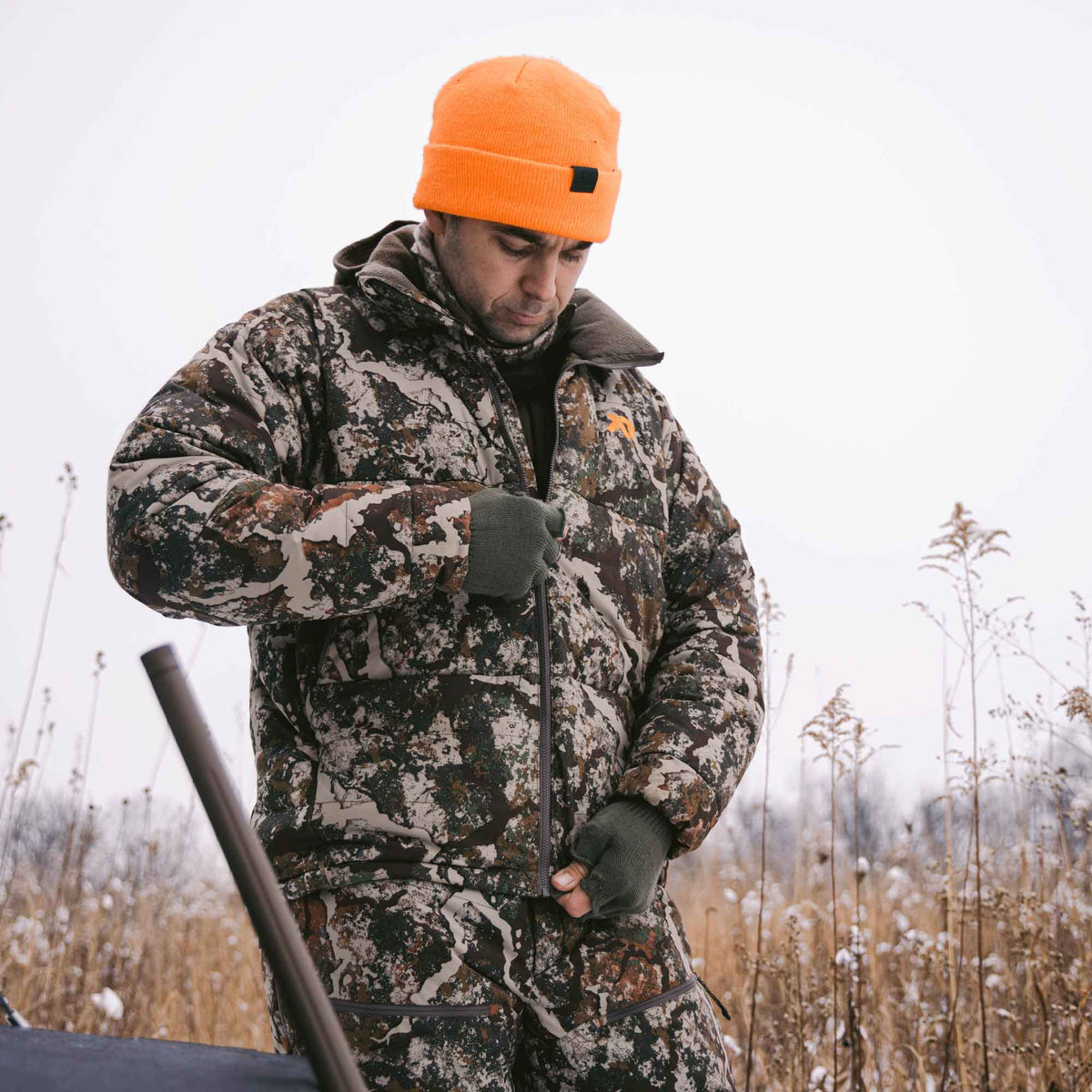 Man zipping up his camo jacket, also wearing an orange beanie and dark green gloves.