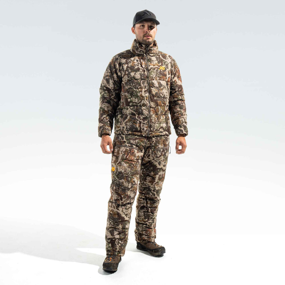 Man wearing a camo jacket and camo pants, a FIRST LITE black cap and brown boots.