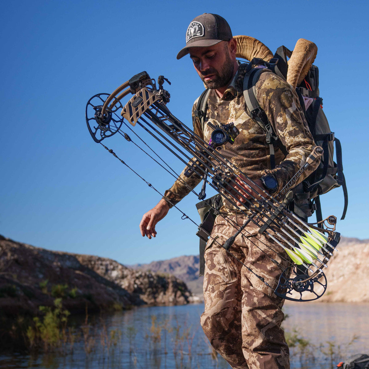 A man in brown and tan camouflage holds a tan and black compound bow with neon yellow-fletched arrows, a dark backpack with large ram horns visible behind him, by a lake.
