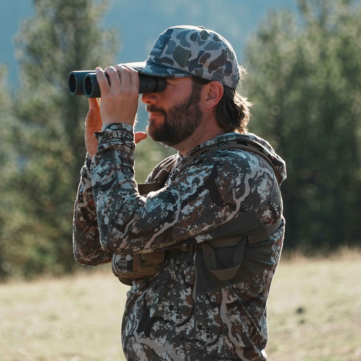 A man in a white, brown, and green camouflage hoodie, a green camo HUSH cap, and a dark binocular chest harness looks through black binoculars.