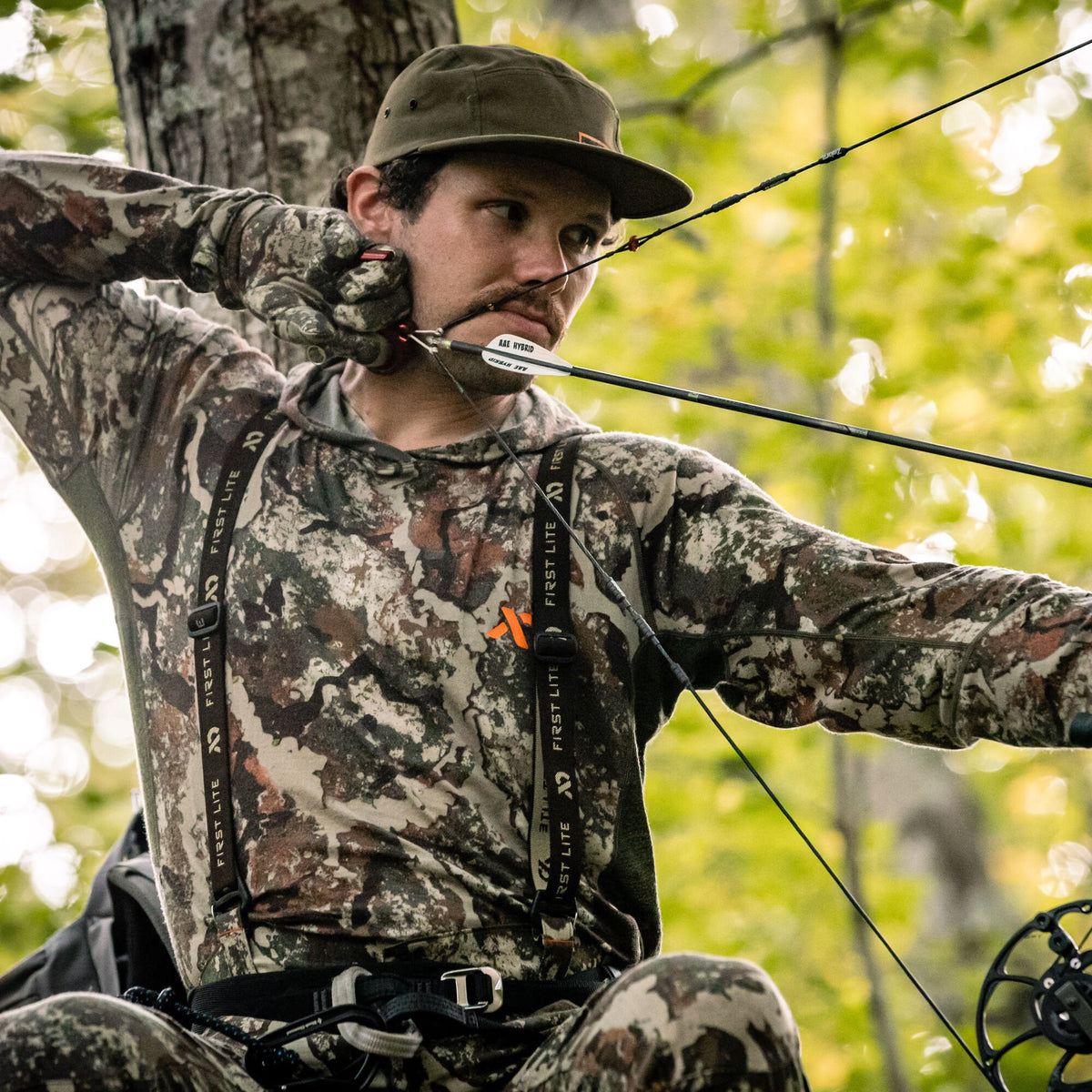 A man in First Lite camouflage hoodie, pants, and gloves, with an olive cap, aims an AAE Hybrid arrow with a compound bow.