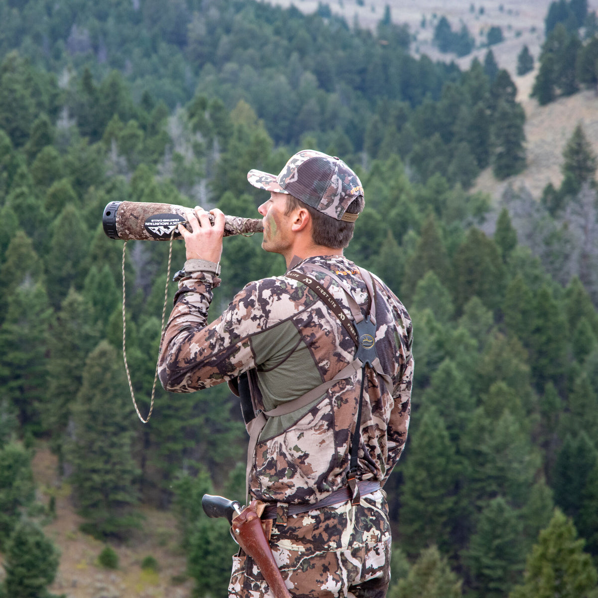 Camouflage Rocky Mountain Hunting Calls call with black cap and lanyard, being used by a hunter. Wearing a brown, cream, and green camo pattern with a mesh hat with First Lite logo on back. Wearing First Lite Suspsenders.