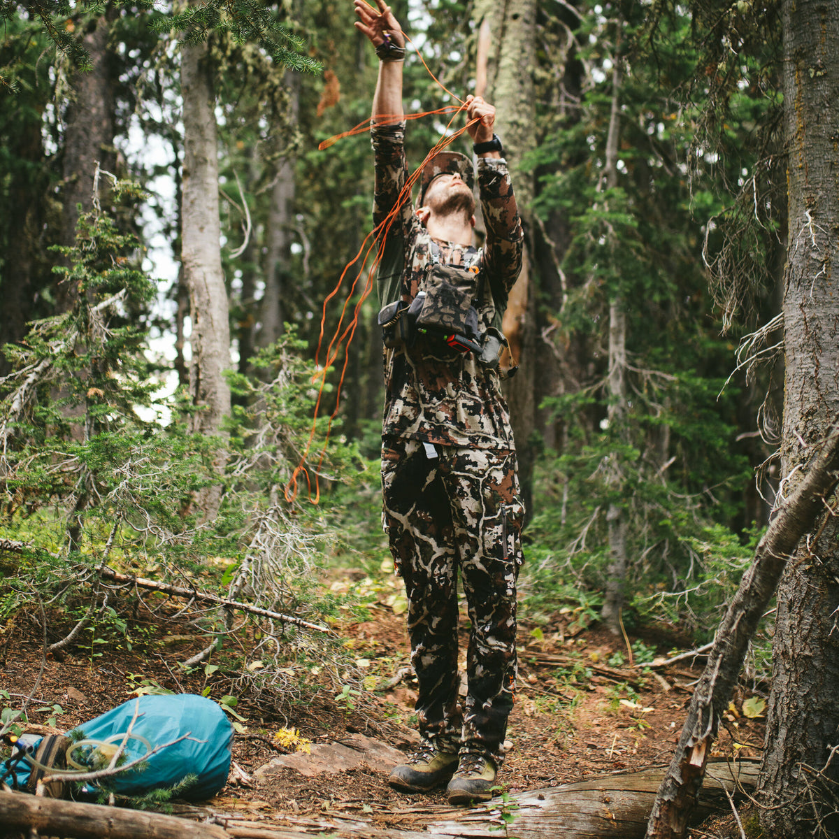 Earth-toned camouflage hunting suit worn by a person with a matching chest rig, reaching upward in a forest.