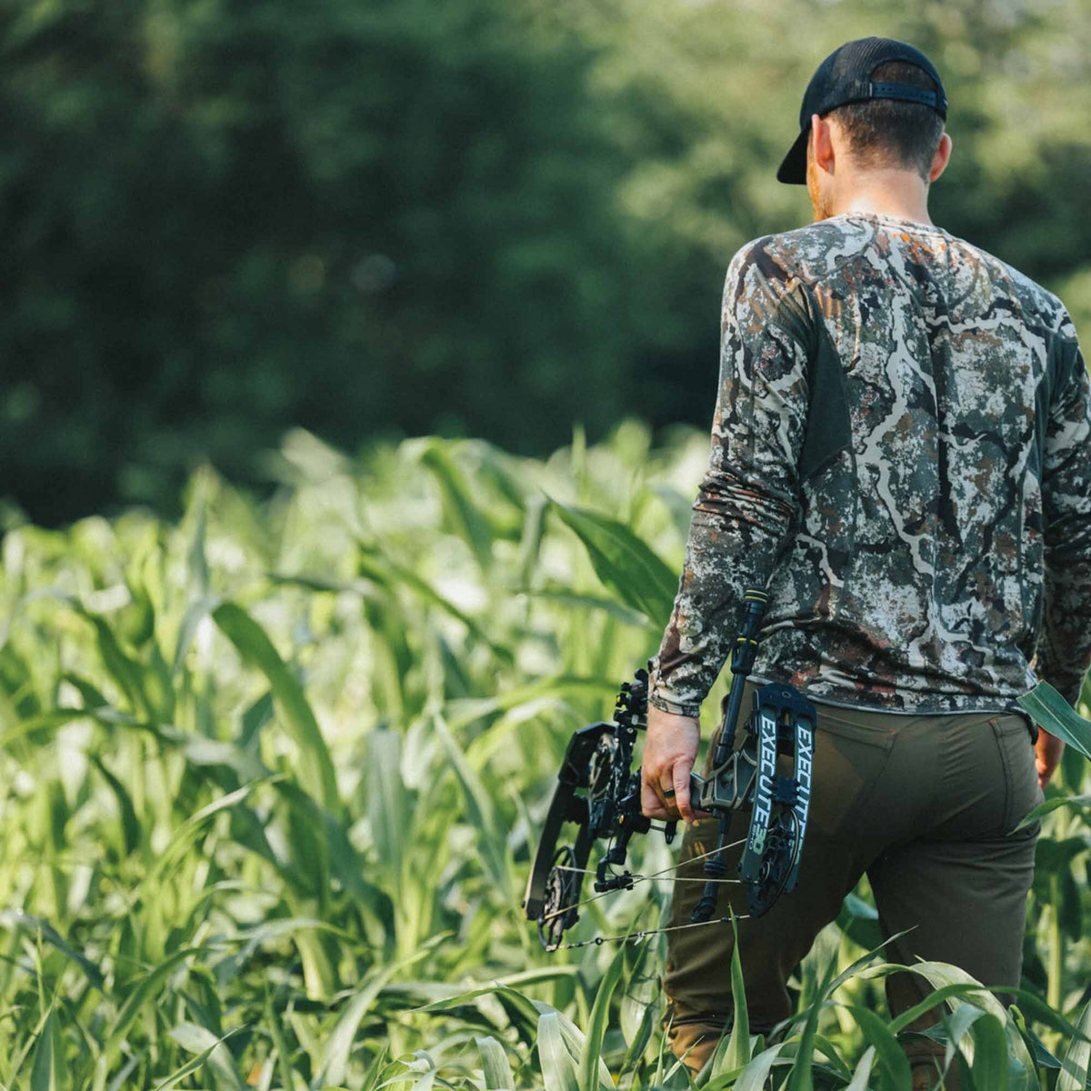 A man in a camouflage long-sleeve shirt and dark olive pants holds a black compound bow with "EXECUTE" branding, walking through a green field.