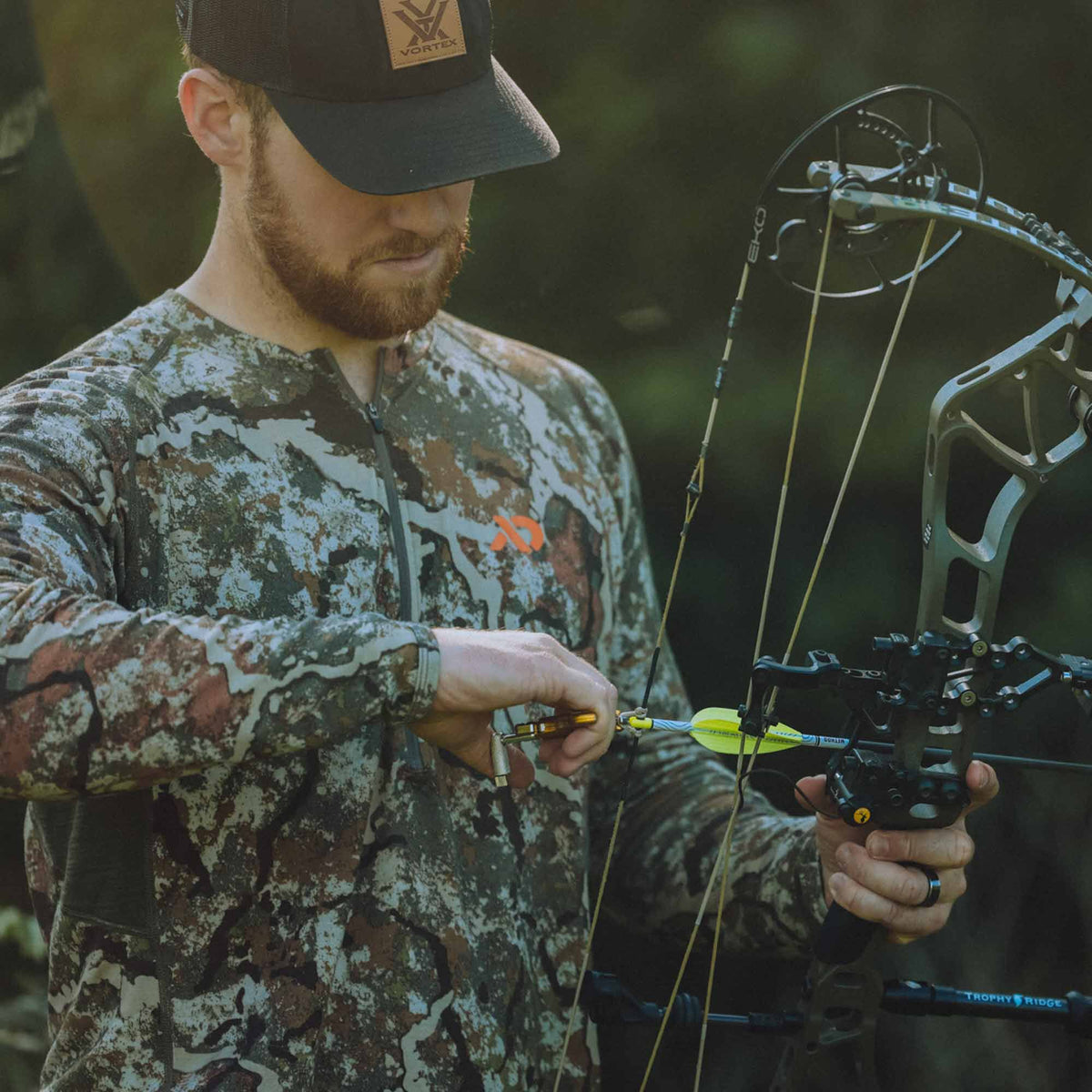 A man in camouflage readies a yellow arrow on a grey compound bow, holding a gold release aid.