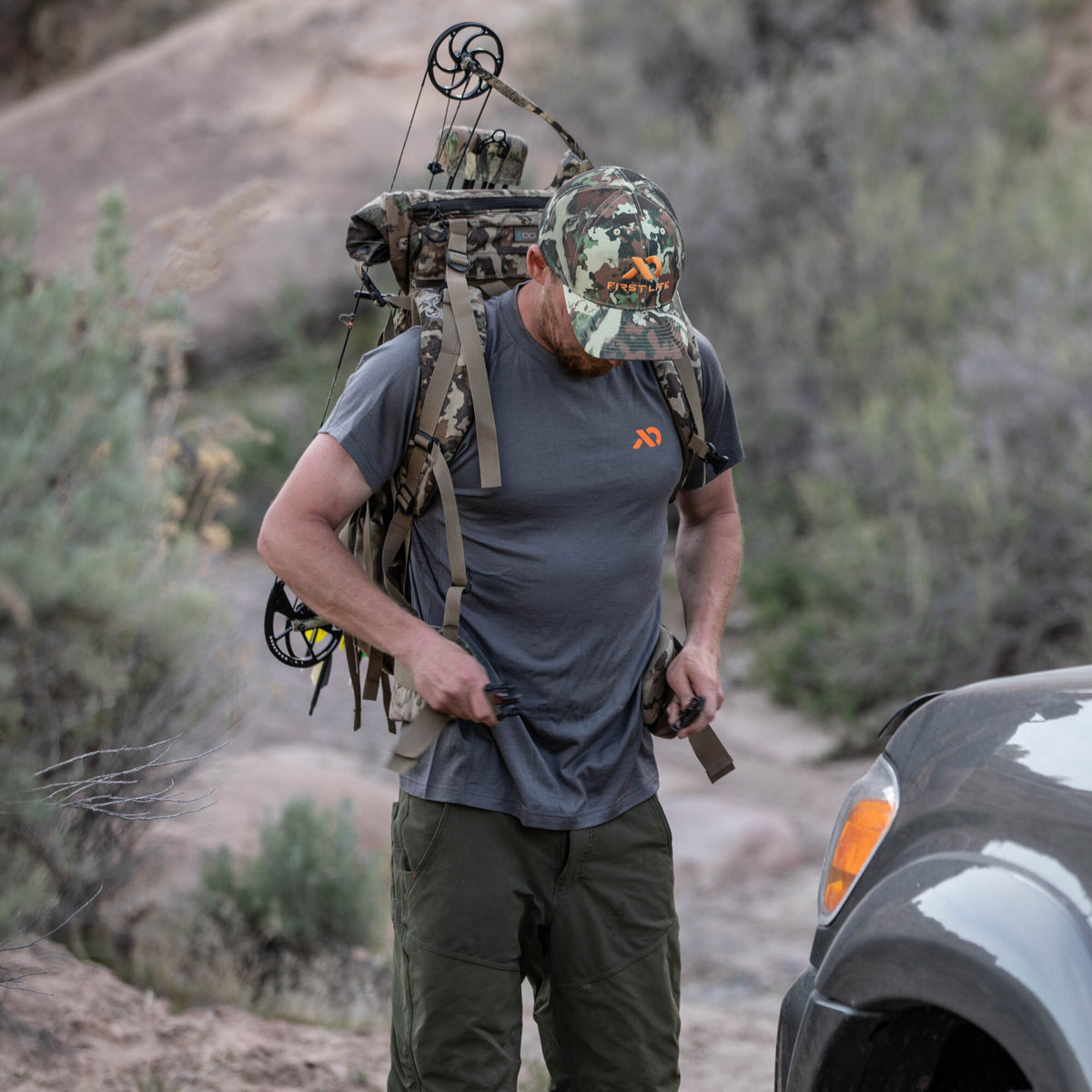 A man wearing a First Lite camo hat and grey shirt adjusts the straps of a large First Lite camo hunting backpack carrying a compound bow.