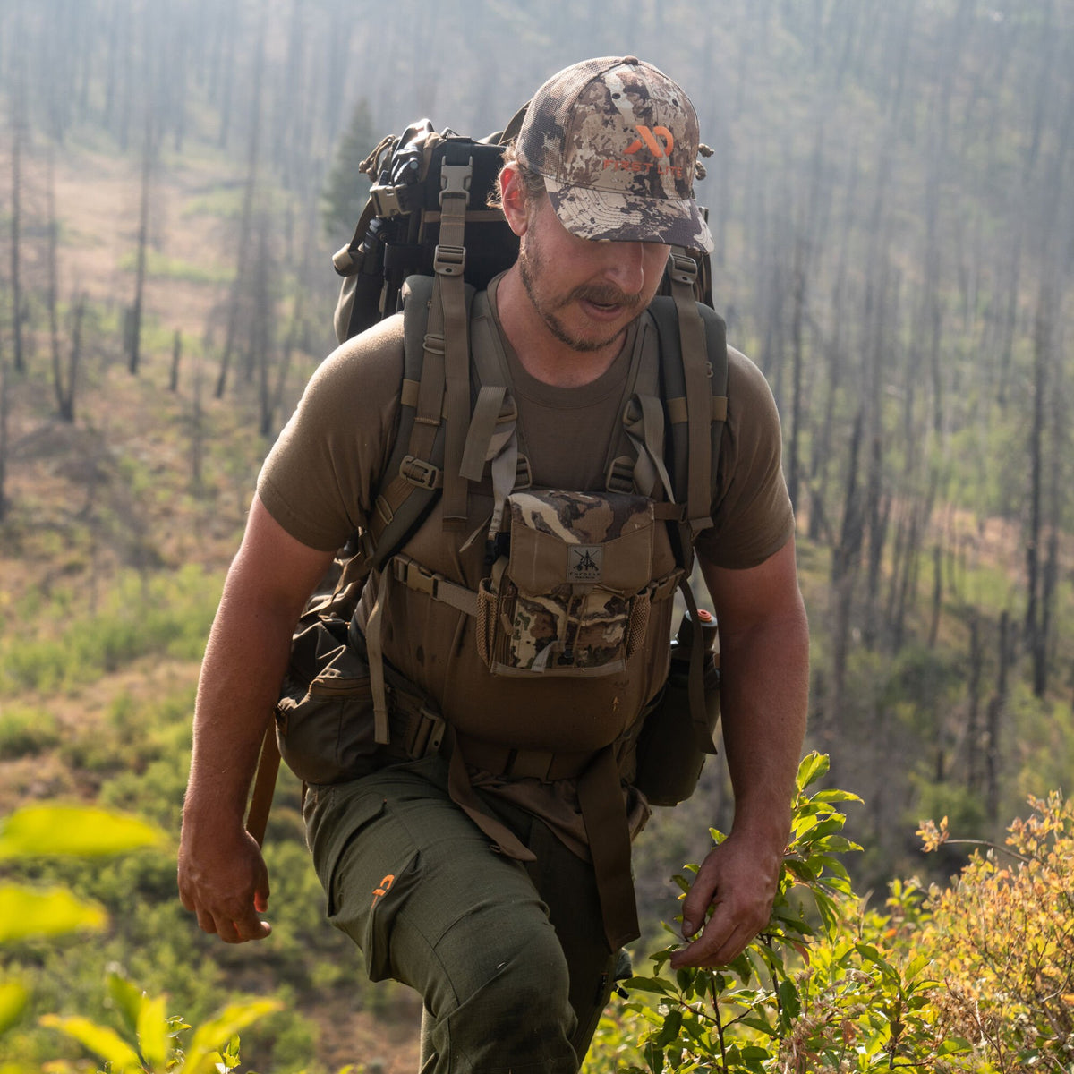Man in camouflage trucker cap and binocular chest rig, olive shirt and cargo pants with orange logos, carrying a large olive tactical backpack.