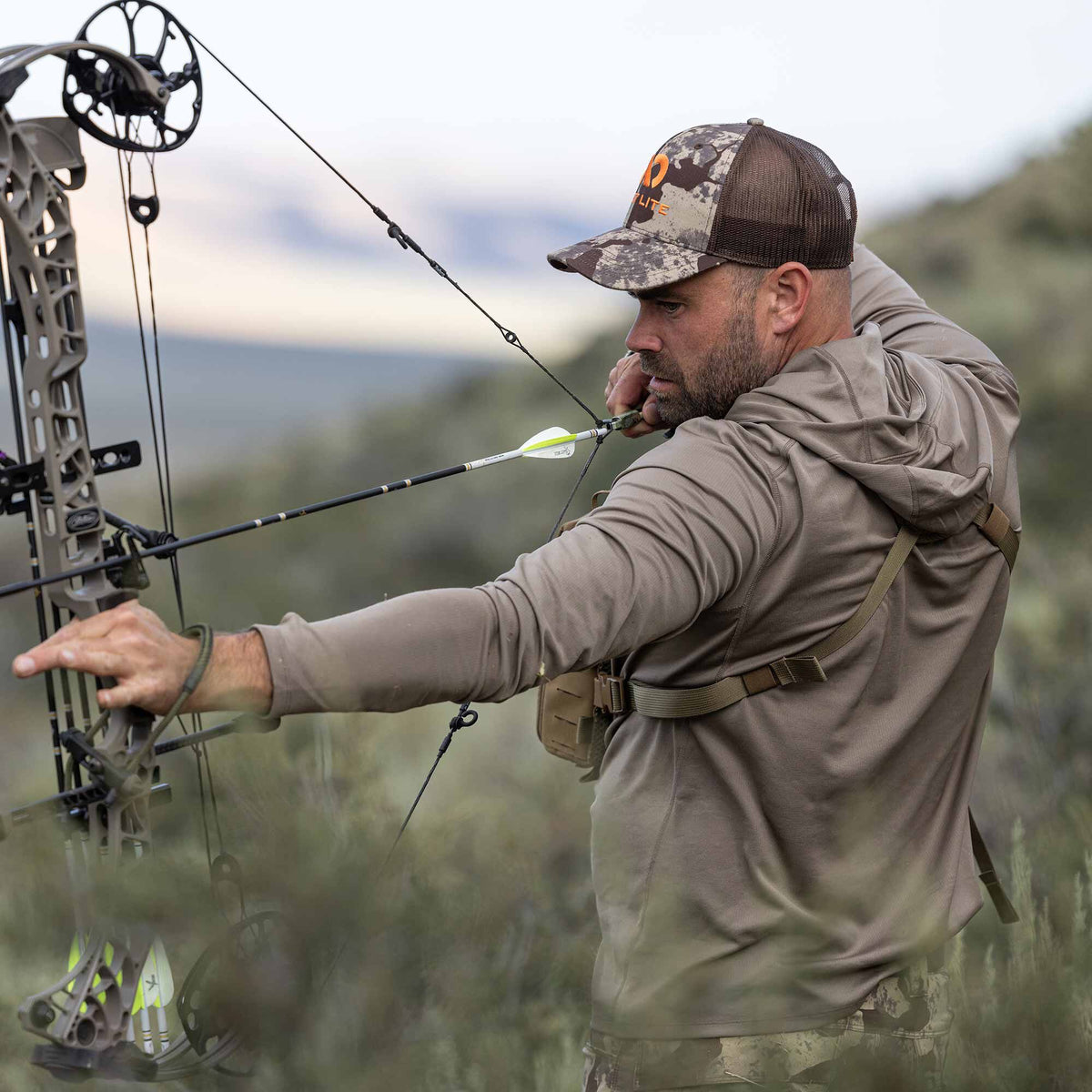 Man drawing a coyote brown compound bow with a black and white fletched arrow, wearing a camouflage cap and a walnut hooded shirt.