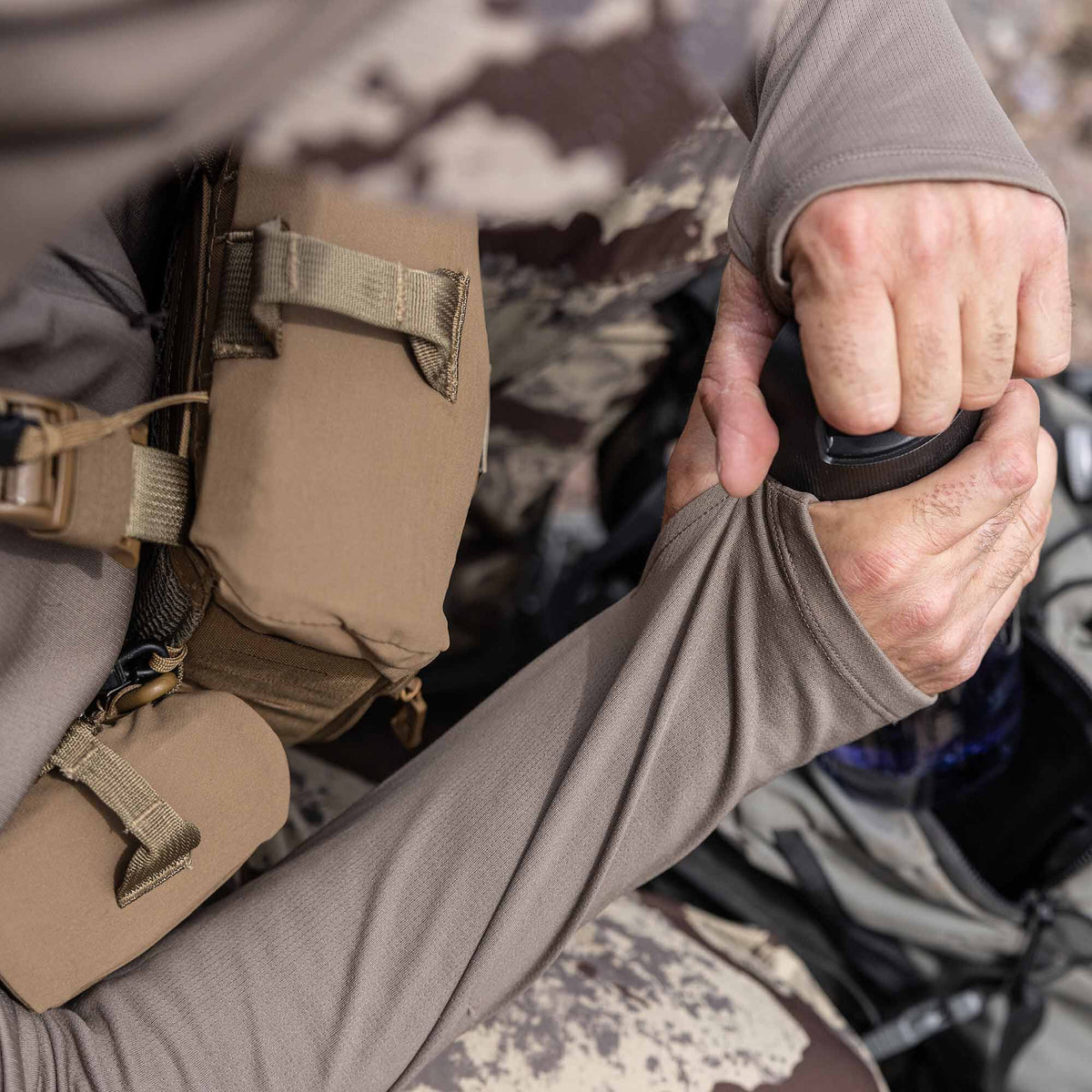 A person wears a Walnut long-sleeved shirt with thumbholes and a subtle grid texture, alongside a tan chest rig.