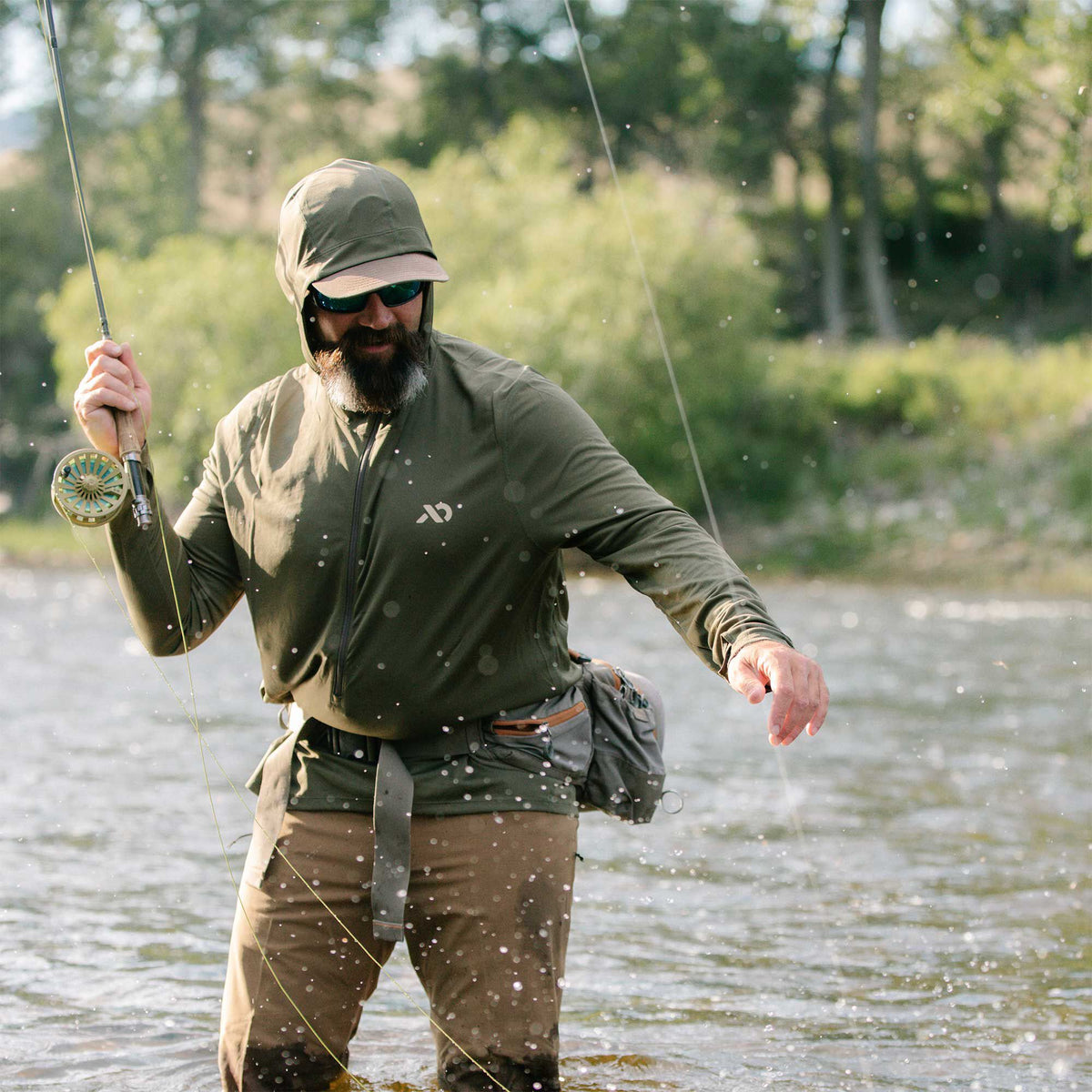 An olive green full-zip hooded long-sleeve fishing jacket with a white chest logo, worn by a bearded man fly fishing in a river.