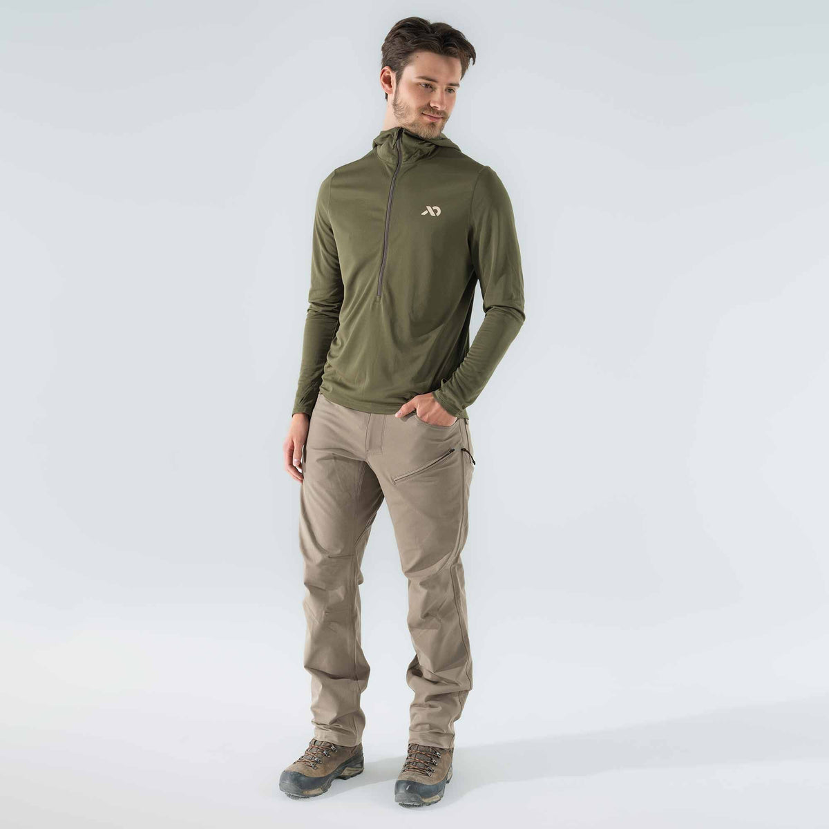 A man wears an olive green quarter-zip hooded long-sleeve shirt with a logo and beige outdoor pants with zippered thigh pockets.