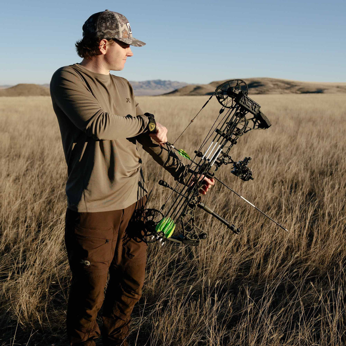 A man wearing a walnut long-sleeve outdoor shirt, brown pants, and a camouflage hat holds a black compound bow with green-fletched arrows.