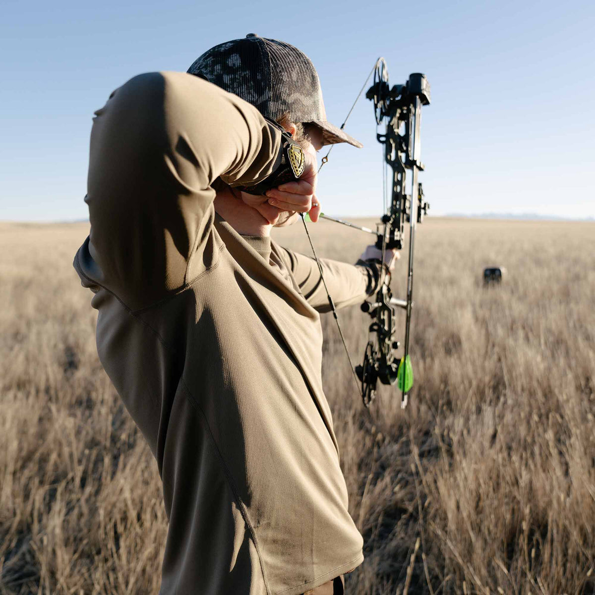 Person in a walnut shirt and camo hat aiming a black compound bow with a green fletched arrow towards a distant target in a dry field.