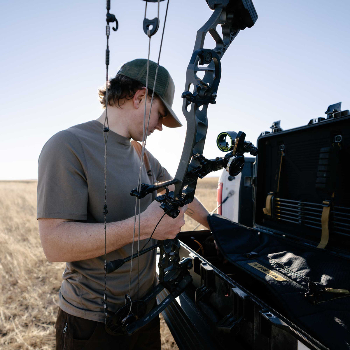 A person takes a black compound bow with a sight and rest out of an open black hard bow case with internal padding and arrow storage.