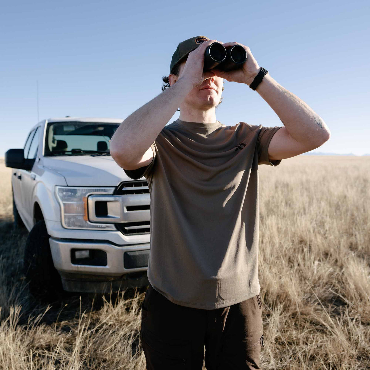 A walnut short-sleeved crew neck shirt with a subtle dark chest logo, worn by a man looking through binoculars in a dry field.
