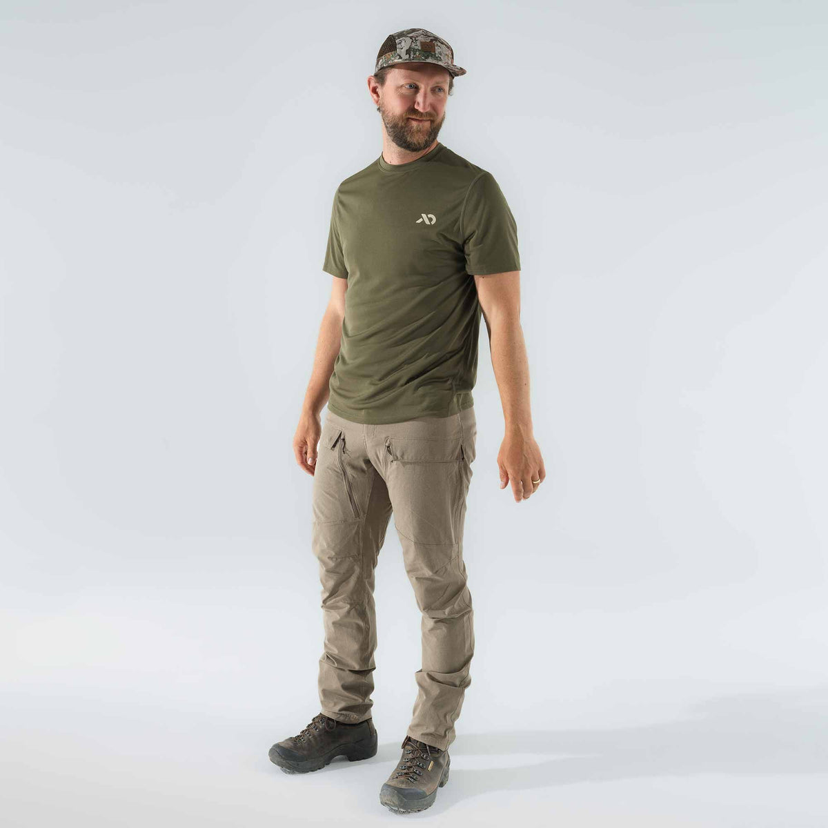 A man wears an olive green crew-neck t-shirt with a light logo, tan technical pants featuring zippered pockets, a camouflage mesh-back hat, and dark brown hiking boots.