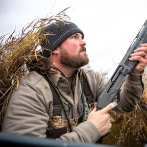 Dark grey semi-auto shotgun held by hunter peering from tall reeds.