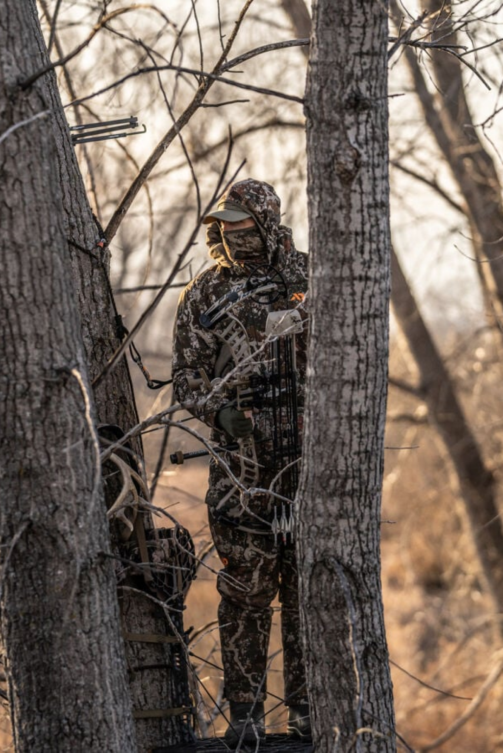 Brown camo hunting suit with mask & cap, worn by hunter holding a bow.