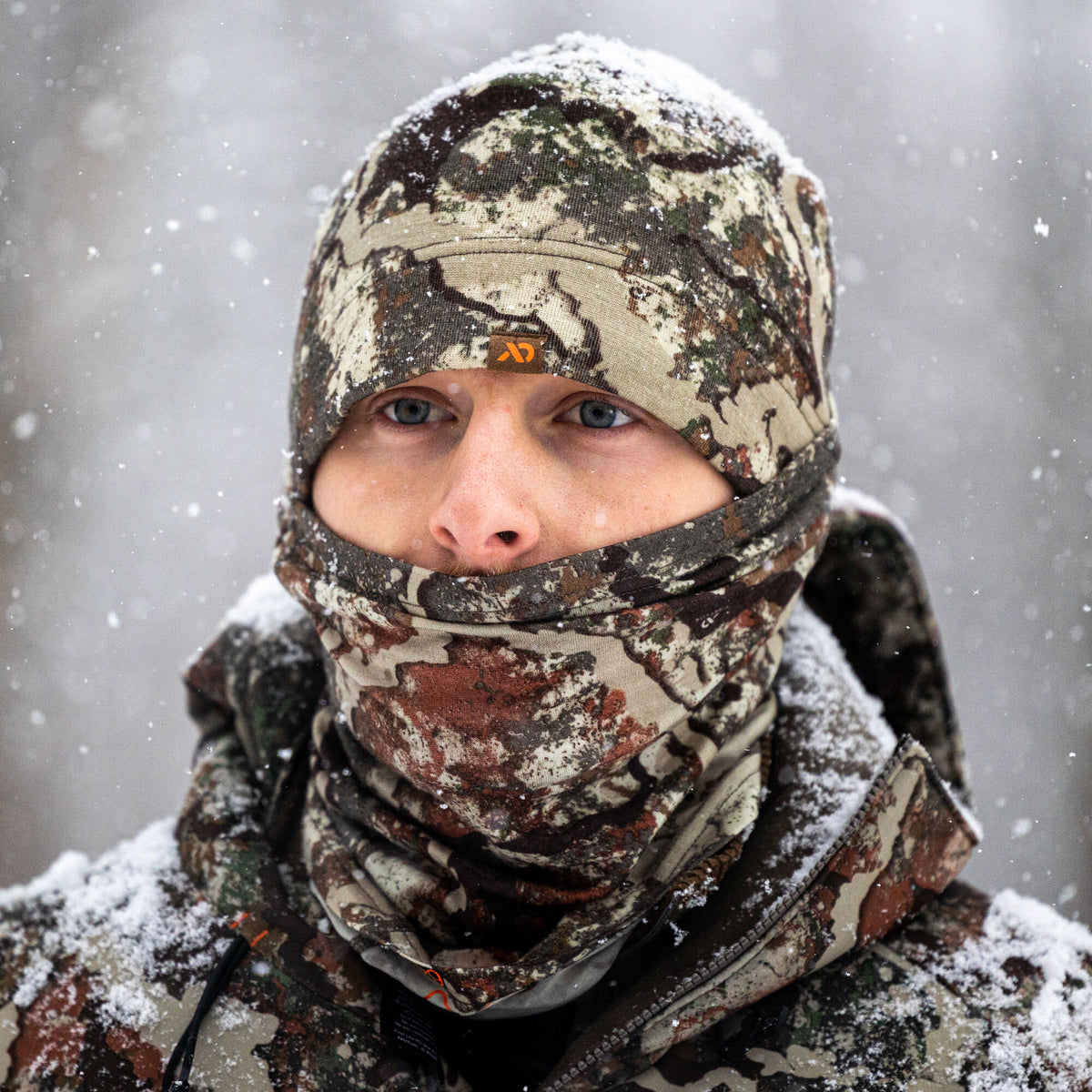 A person wears a brown, tan, and green camouflage balaclava and beanie with an orange logo, lightly covered in snow.