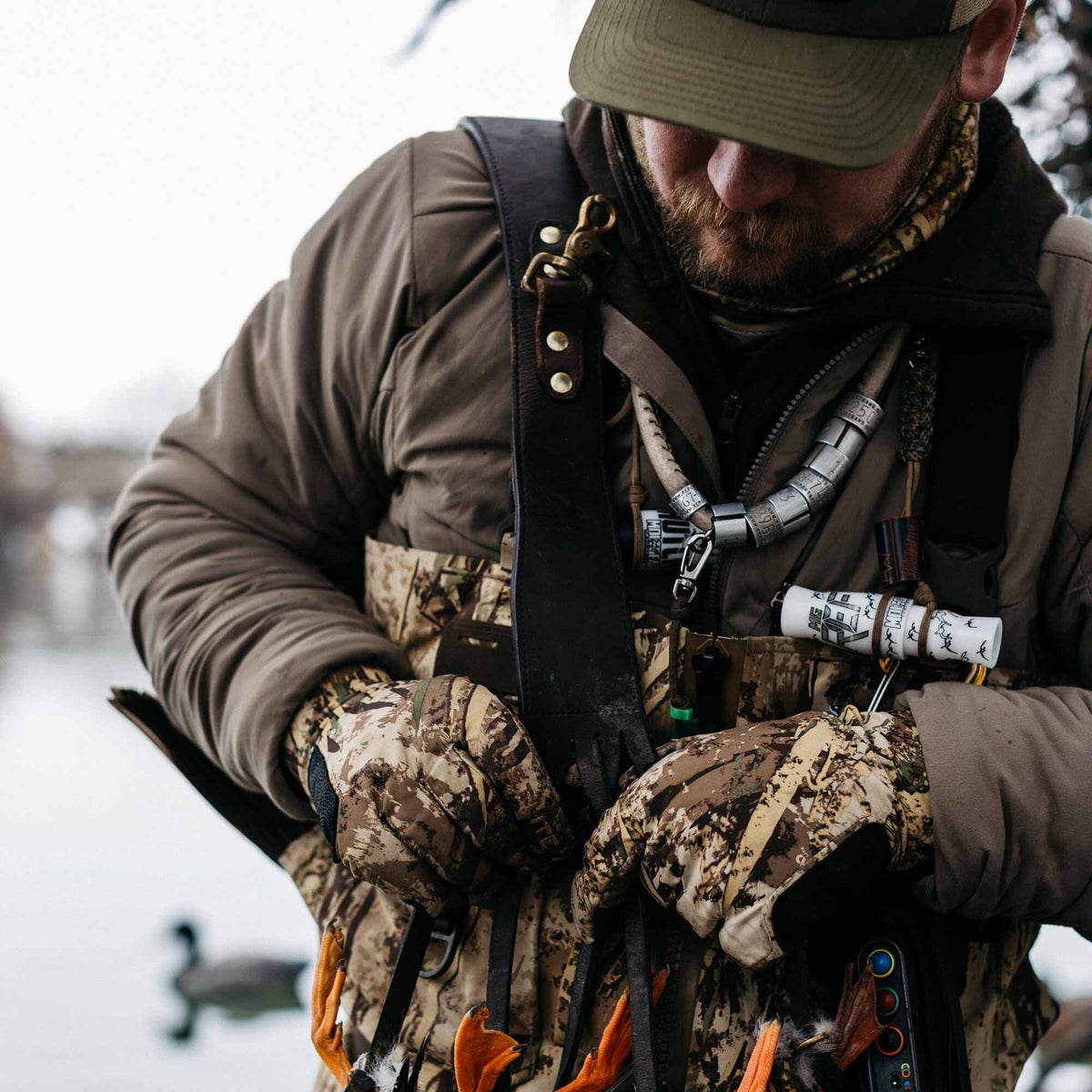 A hunter in an olive jacket and camo gloves wears a dark brown leather lanyard with brass snaps, featuring white duck calls and silver bands, with orange duck feet below.