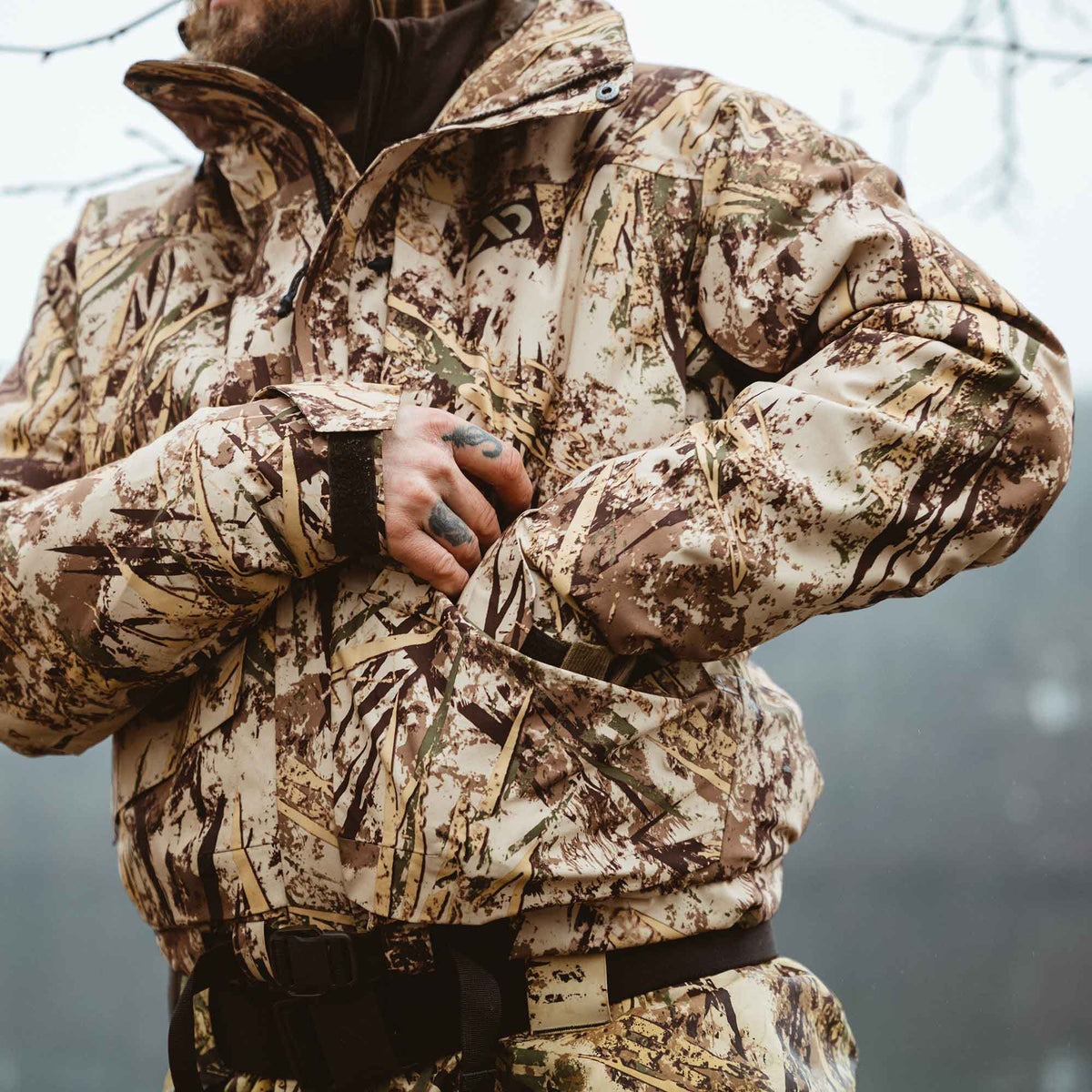 A person wearing a tan, brown, and green dry grass camouflage jacket with a chest logo, reaching into a large front pocket.
