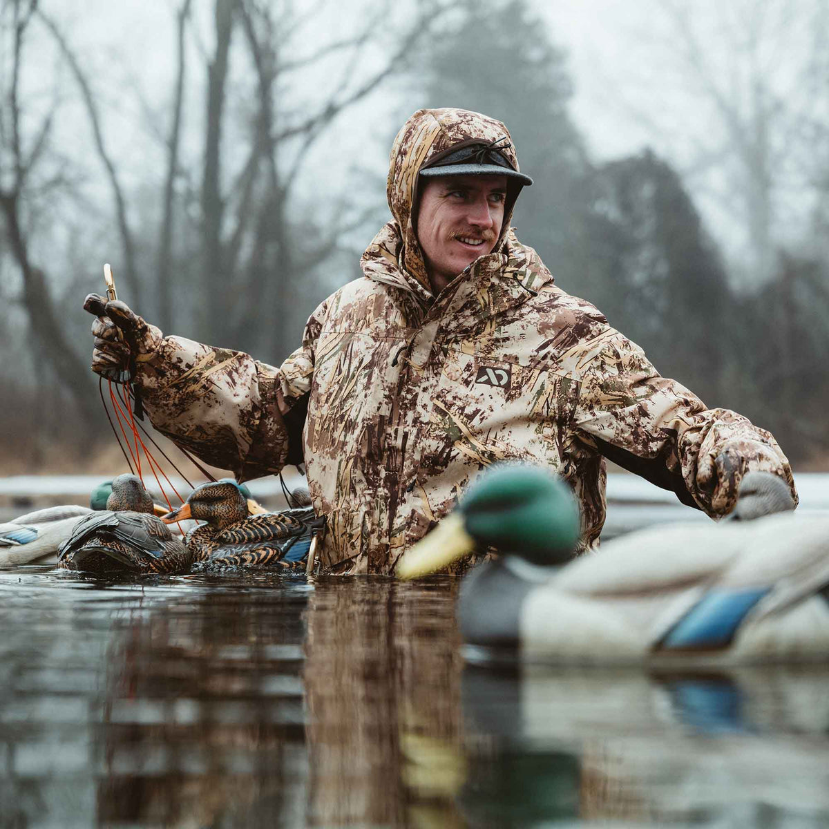A man in a brown, and tan camouflage hooded hunting jacket and matching gloves stands in water surrounded by duck decoys.