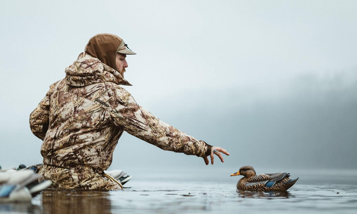 First Lite brown camo hunting suit worn by man reaching for a duck decoy in water.