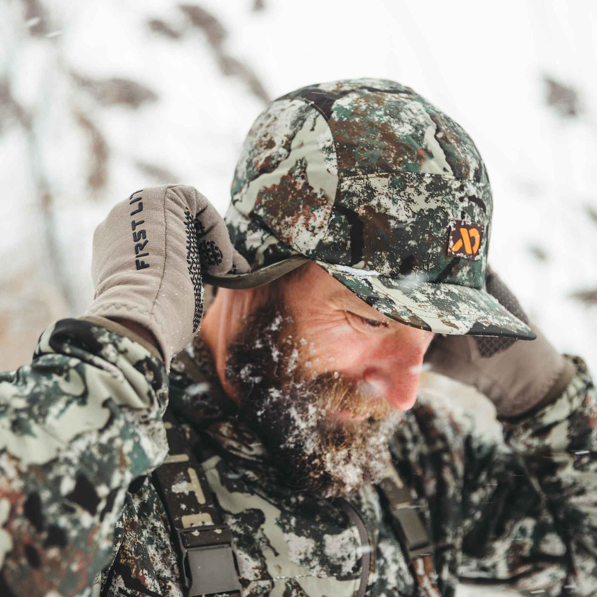 Speckled camouflage cap with integrated ear flaps and an orange logo, worn by a man in a snowy environment.