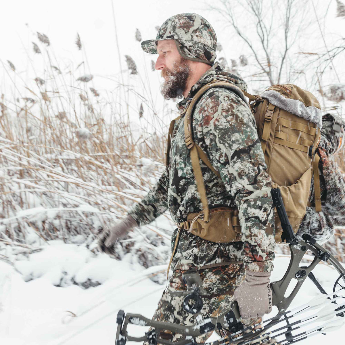 A bearded hunter in snow camouflage walks through snow-covered reeds, carrying a tan backpack with faux fur trim and a compound bow with arrows.