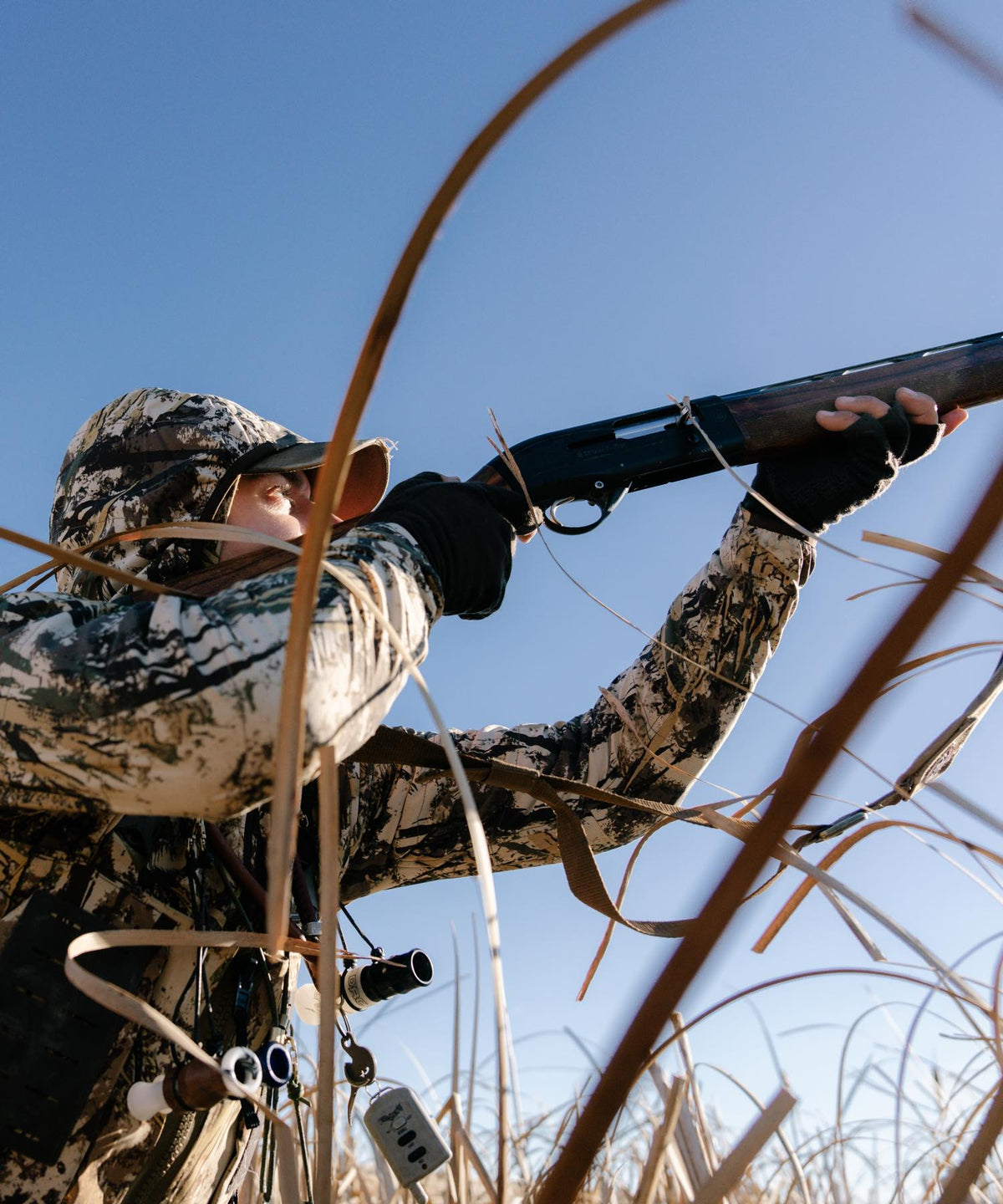 Hunter in white camo aims dark shotgun; fingerless gloves, duck calls, remote visible.
