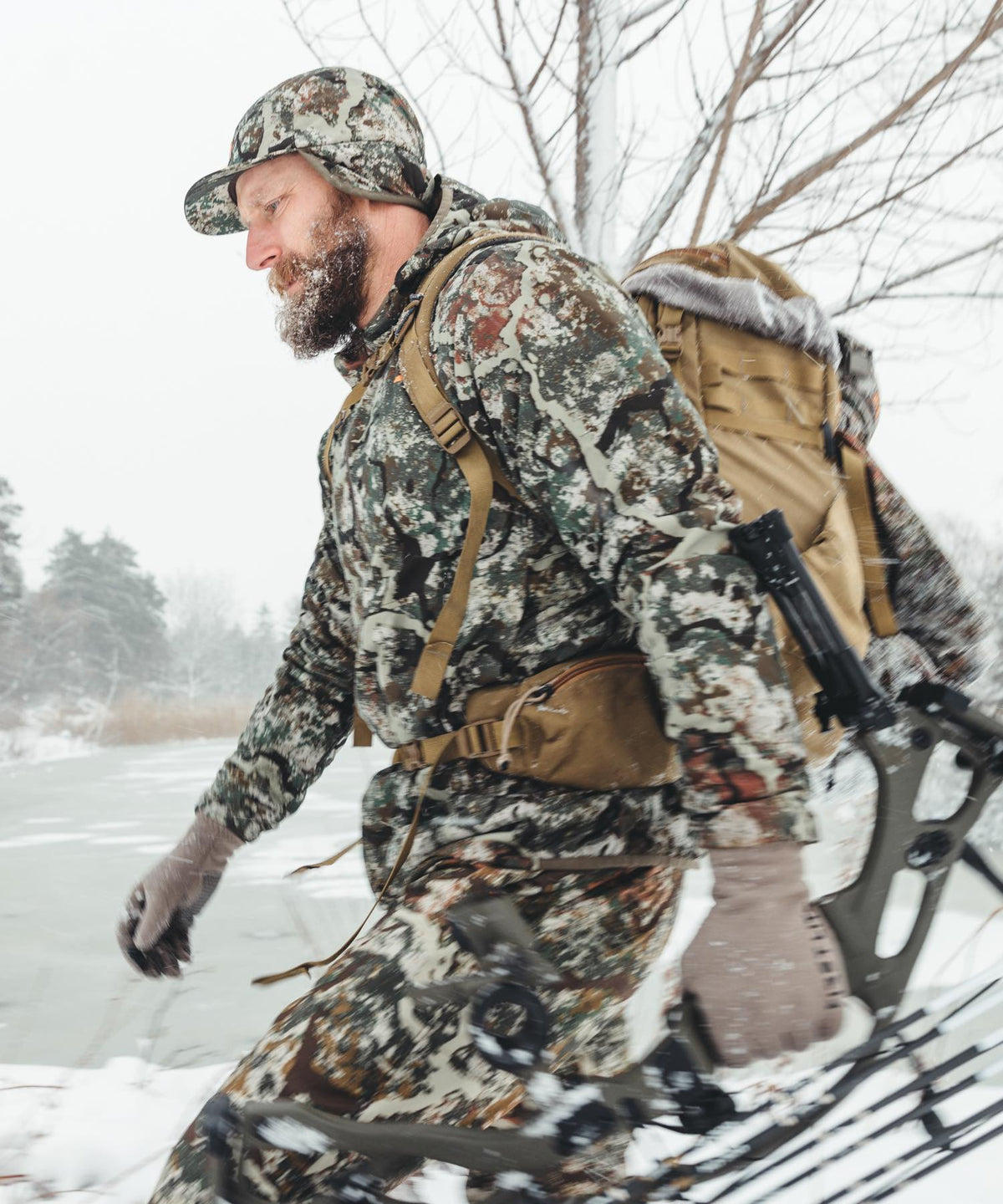Multicam camo hunting suit, tan backpack, and crossbow in snowy environment.