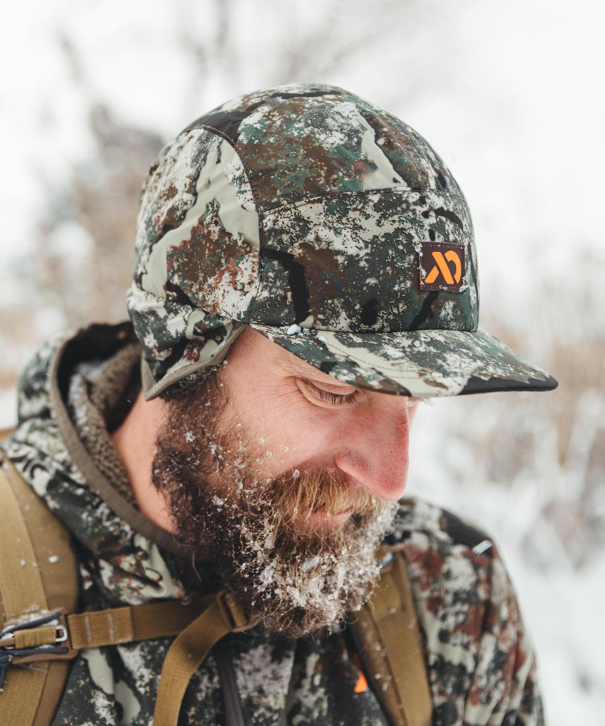 Insulated camo winter cap with ear flaps and orange logo, on man with frosted beard.