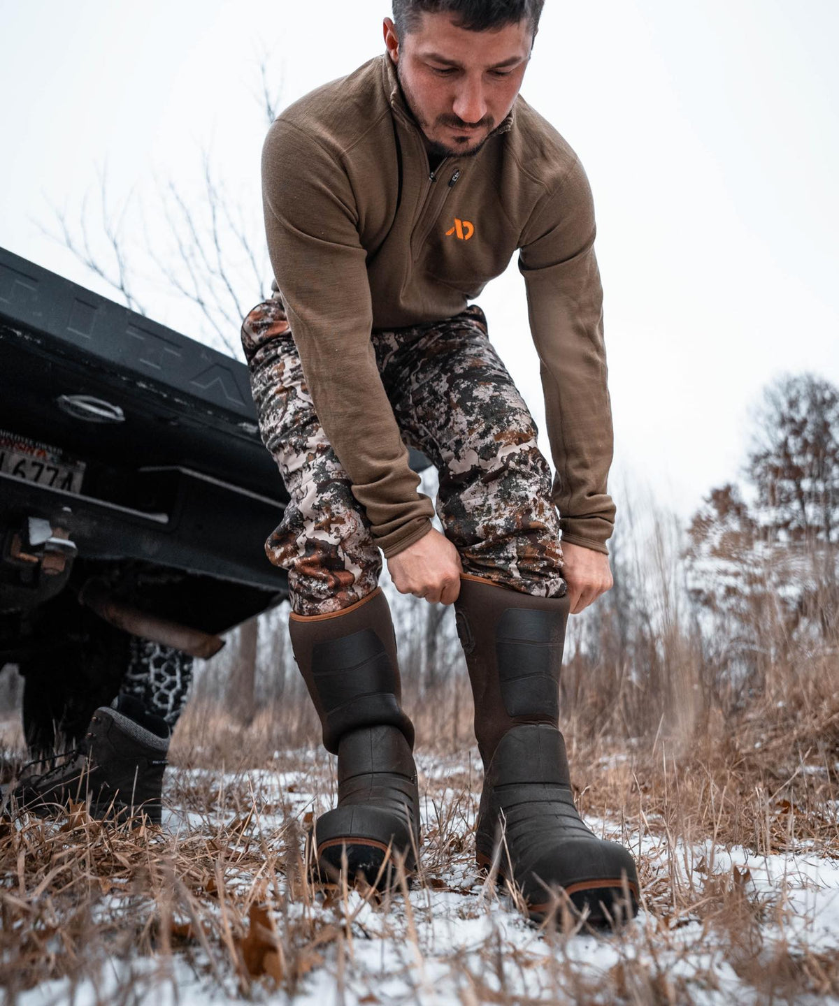 Man pulls on tall dark brown rubber hunting boots with orange trim in snowy grass.