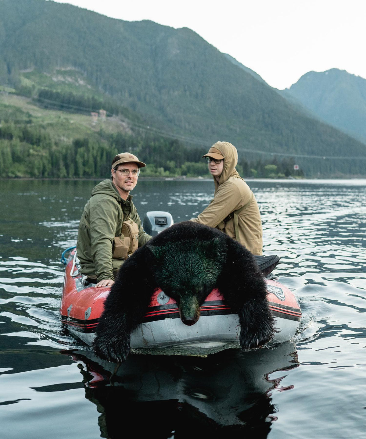Dead black bear in a red-orange inflatable boat with two men on a lake.