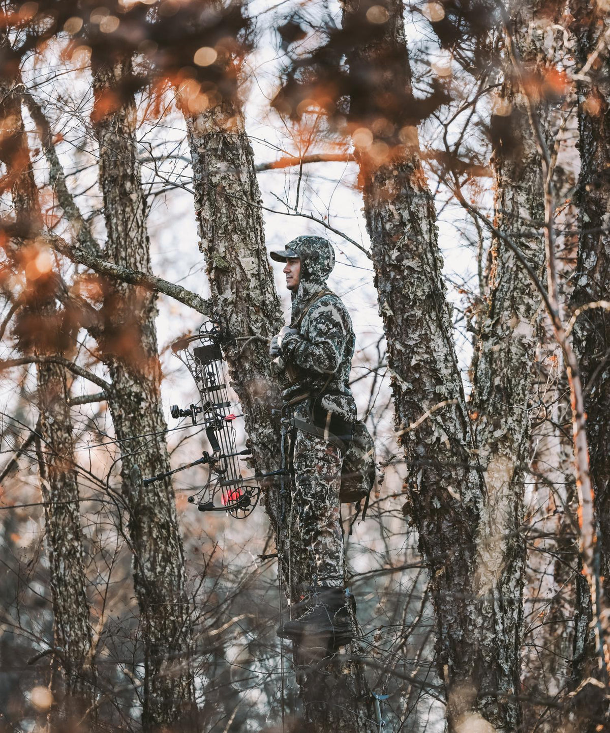 Hunter in white-grey camouflage gear with bow & backpack on a tree stand.