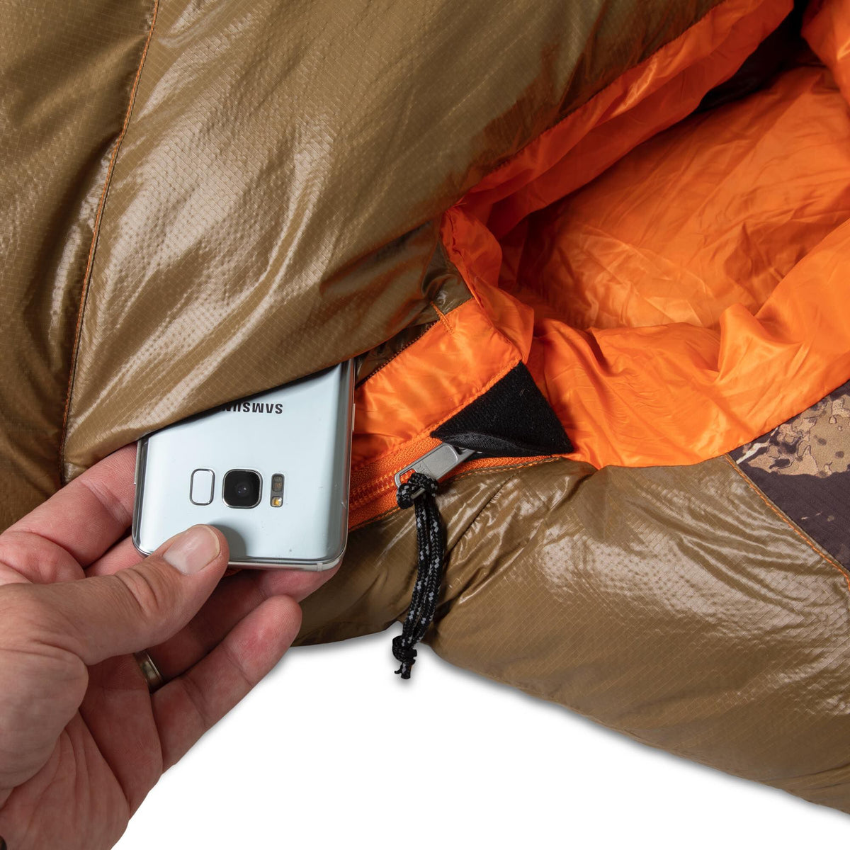 A hand places a silver Samsung phone into the zippered pocket of a brown ripstop sleeping bag with an orange interior.