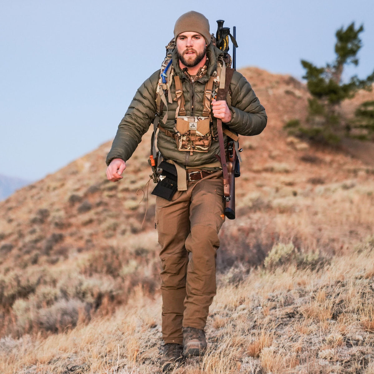 Man in an olive green puffy jacket, brown tactical pants, and tan beanie, with a camouflage chest rig, carrying a rifle and backpack while hiking uphill.