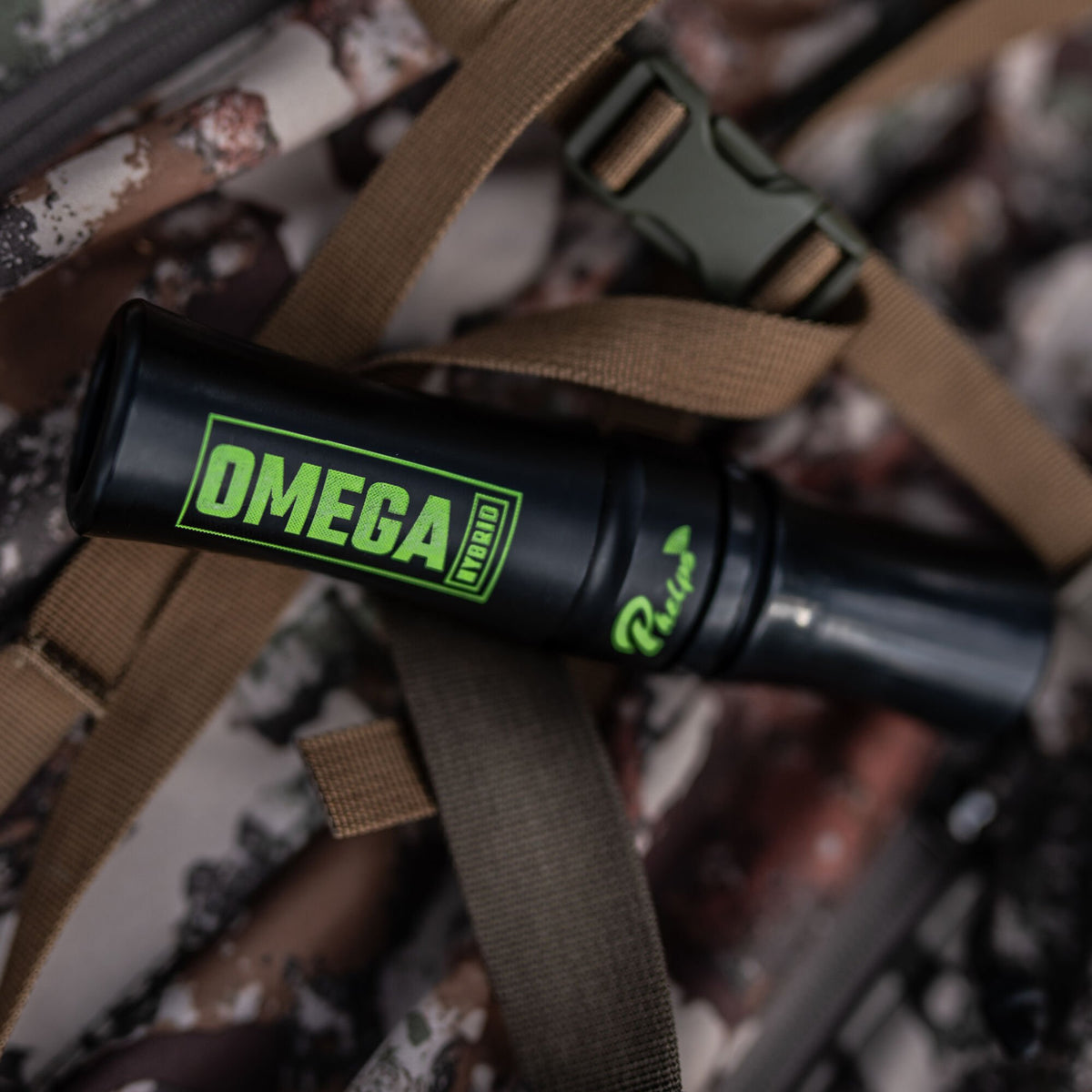 Black game call with green OMEGA HYBRID and Phelps branding, resting on a camouflage backpack.