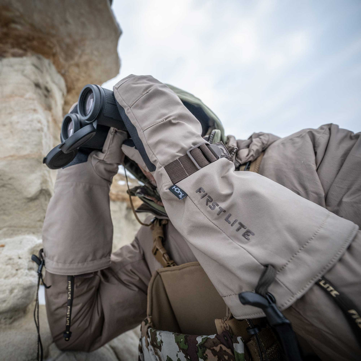 Tan First Lite HDry oversized mitts with an adjustable wrist strap worn by a person looking through binoculars.